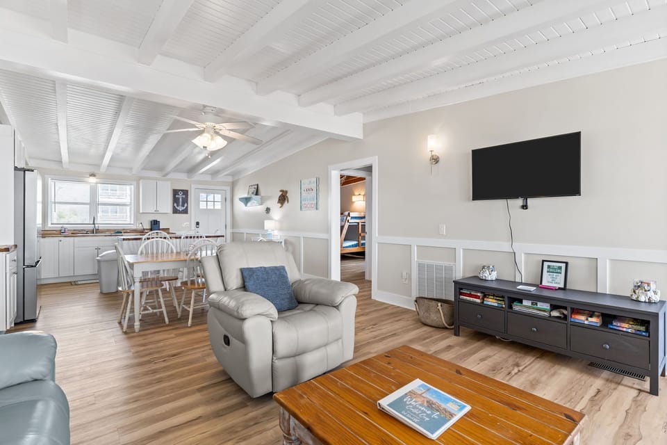 Bright living room with leather sofas, recliner, TV, and coffee table, plus large windows and deck access showcasing beautiful ocean views in this Oak Island, NC beach home.