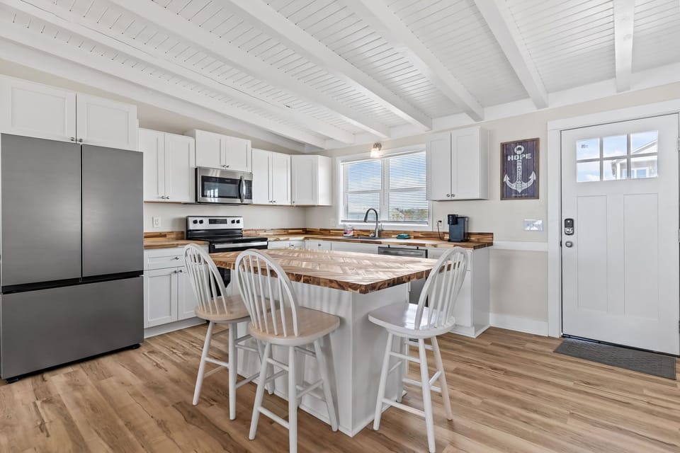 Bright kitchen with white cabinetry, stainless appliances, butcher block counters, and island seating for four, plus coffee maker and window views in this Oak Island, NC beach home.
