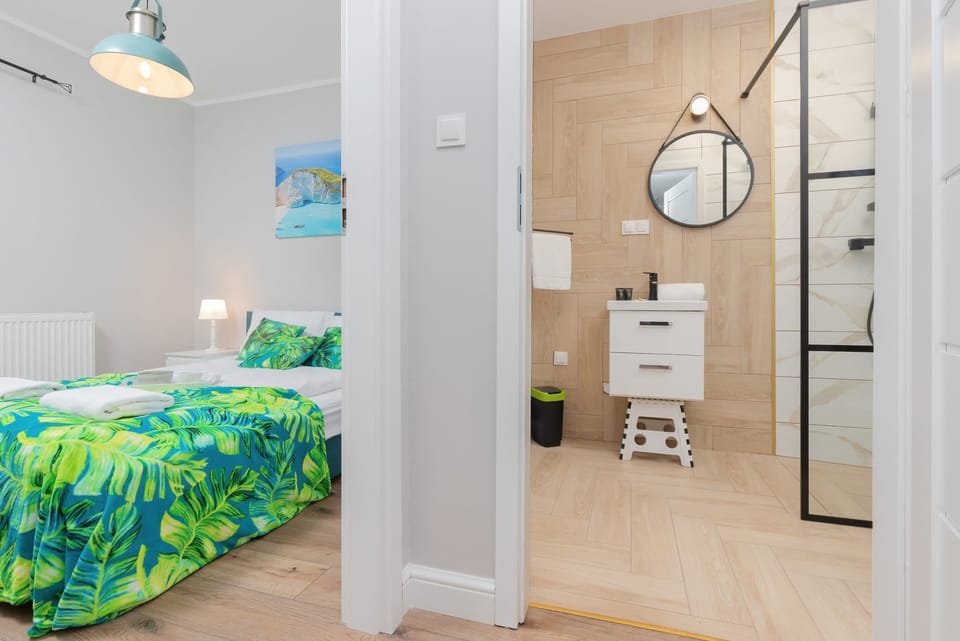 View from the hall to the bedroom and bathroom sink area with a mounted mirror, modern fixtures, and toiletries on the countertop.