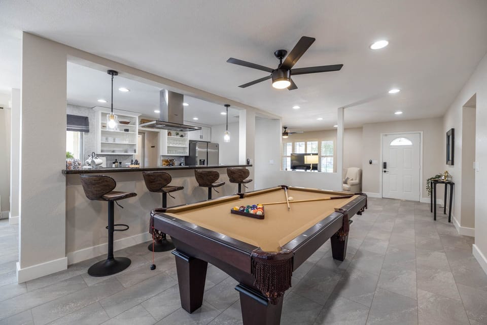 A modern game room with a pool table, a bar area with three stools, a ceiling fan, and an open kitchen in the background. The room is well-lit with recessed lighting.