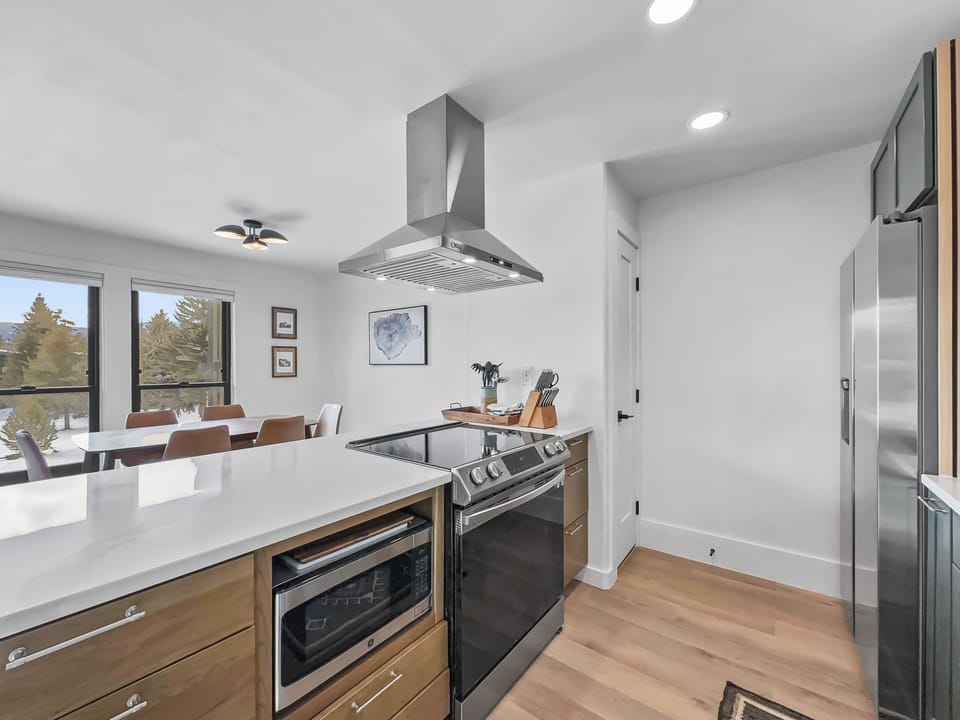 Modern kitchen with a stainless steel stove, range hood, and refrigerator. Light wood cabinets, white countertops, and a dining area in the background with windows overlooking trees.