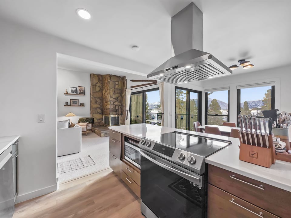 Modern kitchen with stainless steel stove and hood. Adjoining open living area features a stone fireplace and large windows with mountain views. Knife block is on the kitchen counter.