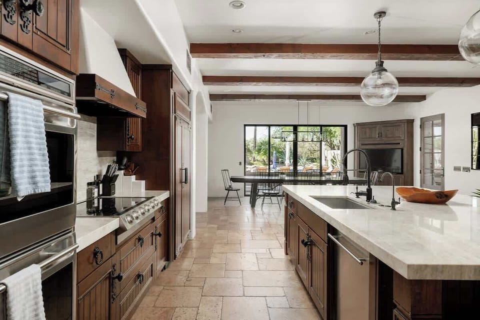 Your gourmet kitchen invites you to cook and gather, with its spacious countertops, top-tier appliances, rustic ceiling beams, and warm natural light streaming in through windows overlooking outdoor greenery.