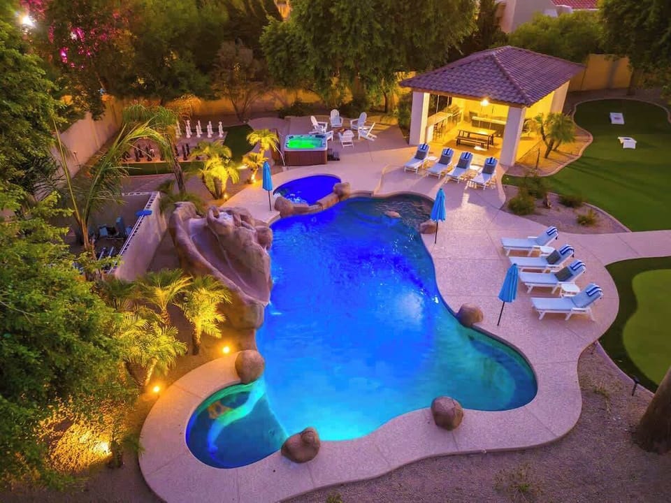Aerial view of resort-style backyard featuring lagoon-shaped pool with waterfall, hot tub, covered outdoor kitchen, and lounge chairs—illuminated at twilight and surrounded by lush landscaping and palm trees.