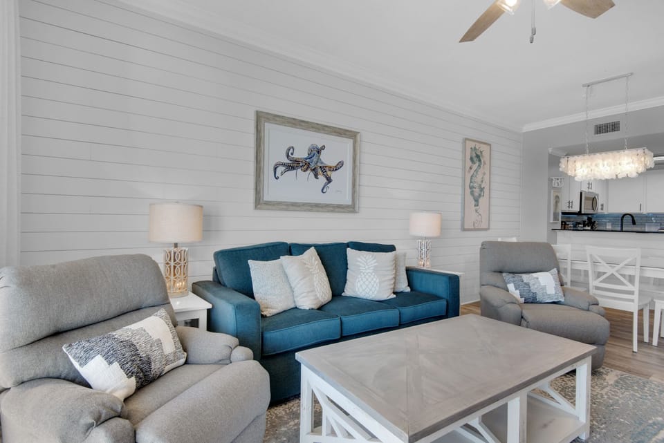 Living Room - Indulge in seaside luxury and comfort with this bright living room, featuring plush furniture including 2 recliners, a large flat-screen TV, and a private balcony over the beach. 
