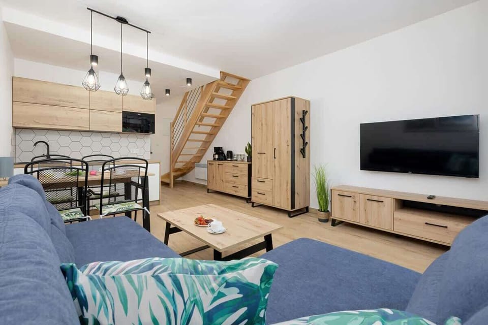 A cozy space with wooden flooring, a large TV mounted on the wall, a light wooden TV stand, and a comfortable navy-blue sofa with a small wooden coffee table. The room is bright, featuring large windows that let in natural light.