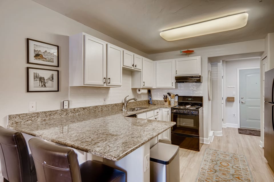 A modern kitchen with white cabinets, granite countertops, a stainless steel sink, black appliances, and two brown cushioned bar stools. Artwork and a fish decoration adorn the walls.