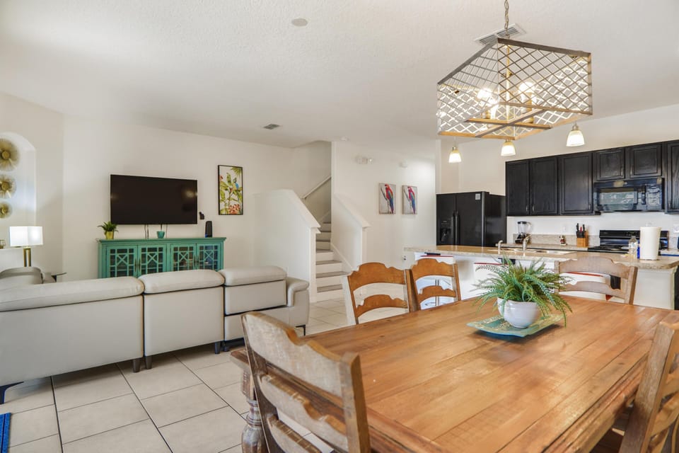 Wood dining table with six chairs sits below a geometric metal light fixture near the open kitchen and living room.