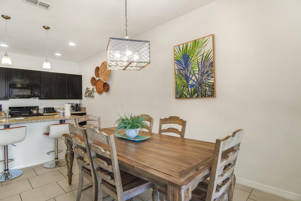 Rustic wooden dining table seats six beneath a metal-framed pendant light and tropical wall art.