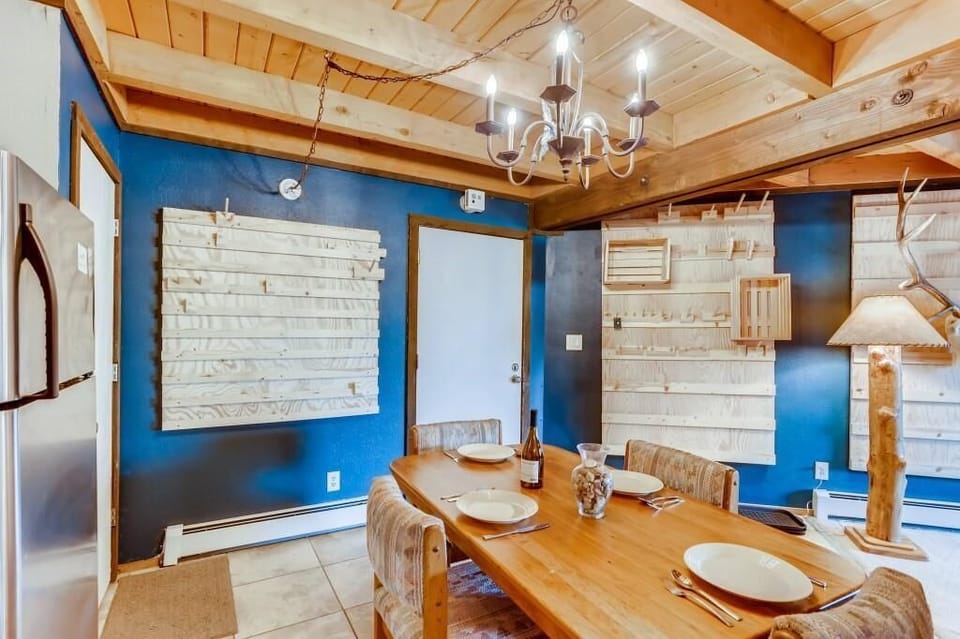 Dining area in a rustic wooden decor with a table set for four. Features a chandelier, wooden ceiling, blue walls with wooden paneling, and a refrigerator on the left.