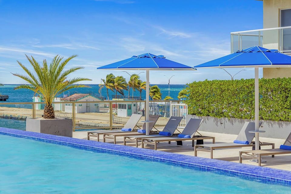 Sparkling pool meets blue sky and ocean horizon, where loungers await beneath vibrant umbrellas and tropical palms sway—a sun-soaked retreat that blends relaxation with breathtaking coastal views.