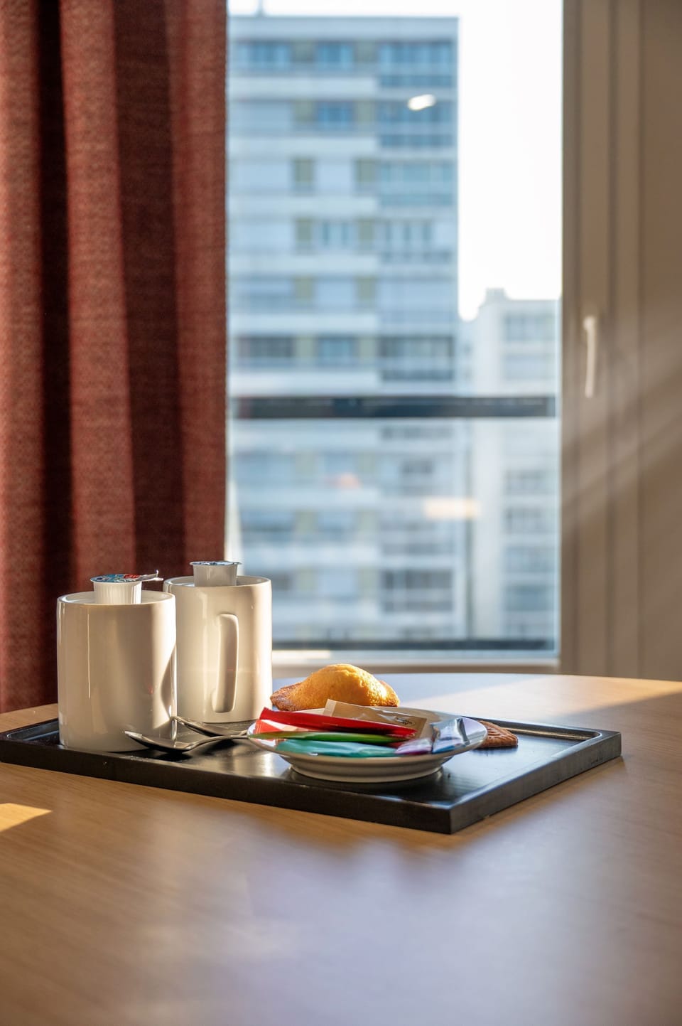 A cozy breakfast tray featuring white mugs, a golden pastry, and colorful packets of condiments, set against a backdrop of a modern building.