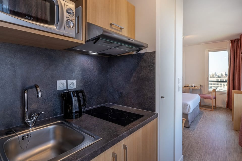 A well-equipped kitchenette displaying stainless steel appliances and a glimpse of the adjoining hotel room.