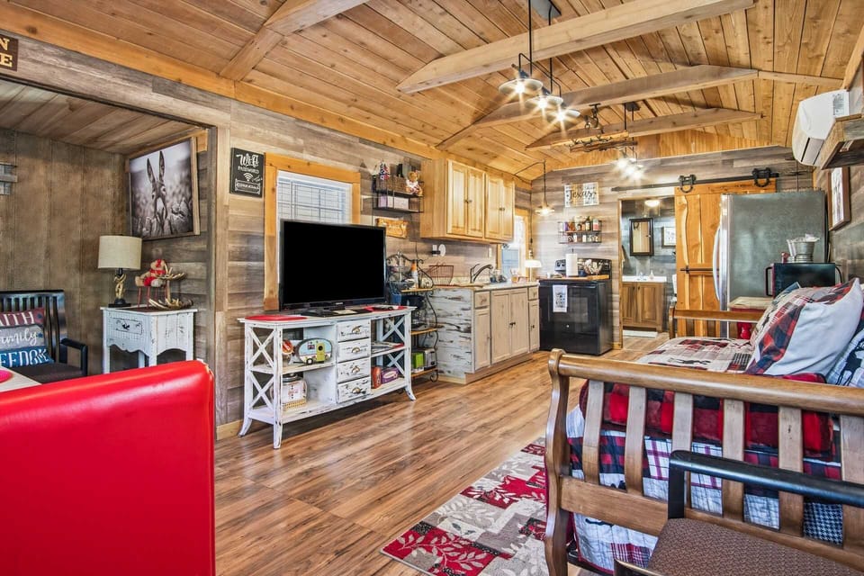 The interior of a cozy, open-concept cabin featuring vaulted wood-plank ceilings, a well-equipped kitchenette, and a living area with a television, red accents, and rustic wooden finishes throughout.