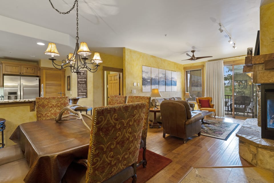 A cozy living and dining area with wooden flooring. It features a dining table, chairs, leather sofas, a fireplace, and kitchen area with stainless steel appliances. Numerous decorations adorn the space.