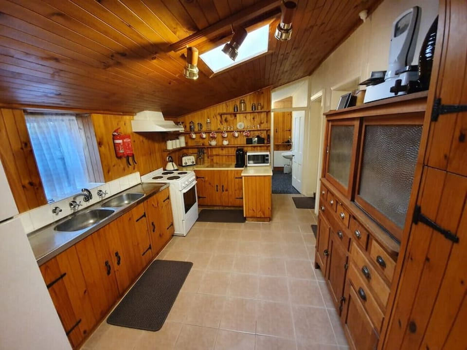 This cozy country-style kitchen features warm wooden paneling on the walls and ceiling, complemented by modern appliances and a skylight that adds natural light. The space is well-equipped with a double sink, electric stove, microwave, coffee machine, and