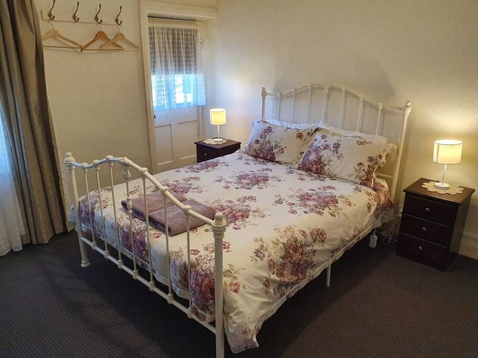 This cozy bedroom features a white metal-framed double bed adorned with floral bedding, two wooden bedside tables with lamps, and a window with sheer curtains for natural light. Wall hooks with hangers offer convenient storage, adding to the room’s charmi
