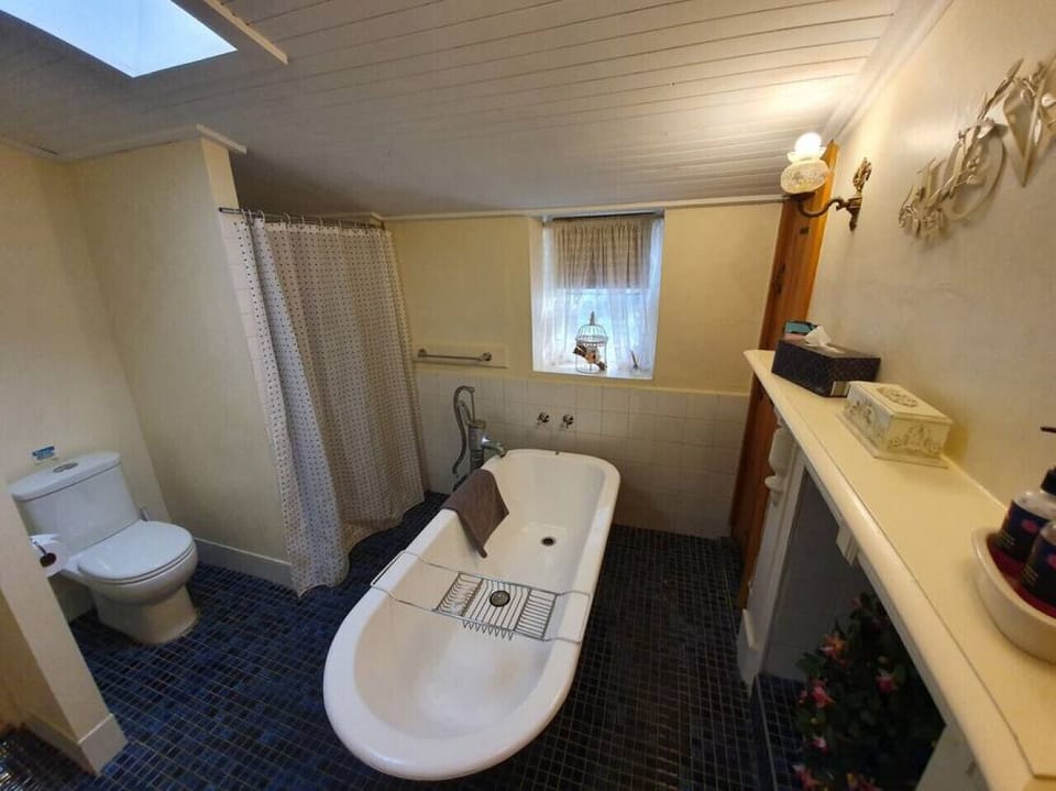 This charming bathroom features a classic freestanding bathtub, a modern toilet, and a shower area with a privacy curtain. A small window with sheer curtains allows natural light to brighten the space, while decorative touches and vintage-style fixtures a