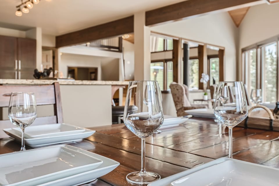 Close-up view of a modern dining table set with square white plates and wine glasses in a bright, open-concept living and dining area with large windows and wooden beams.