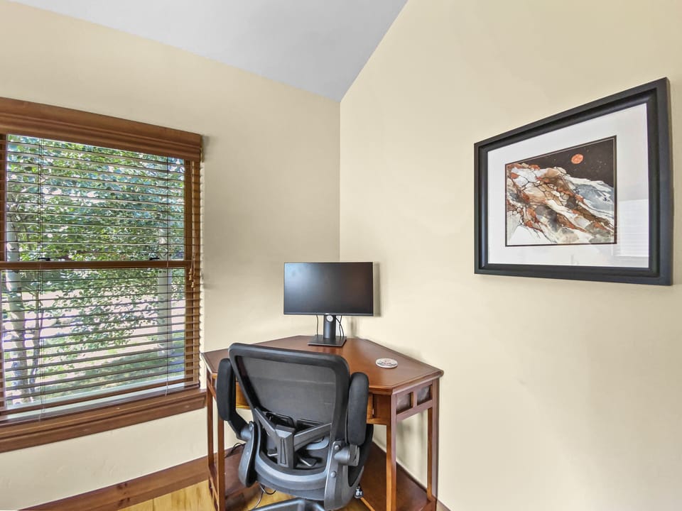 A small home office setup with a desk, chair, computer monitor, and mouse. A window with blinds and a framed picture on the wall are also visible.