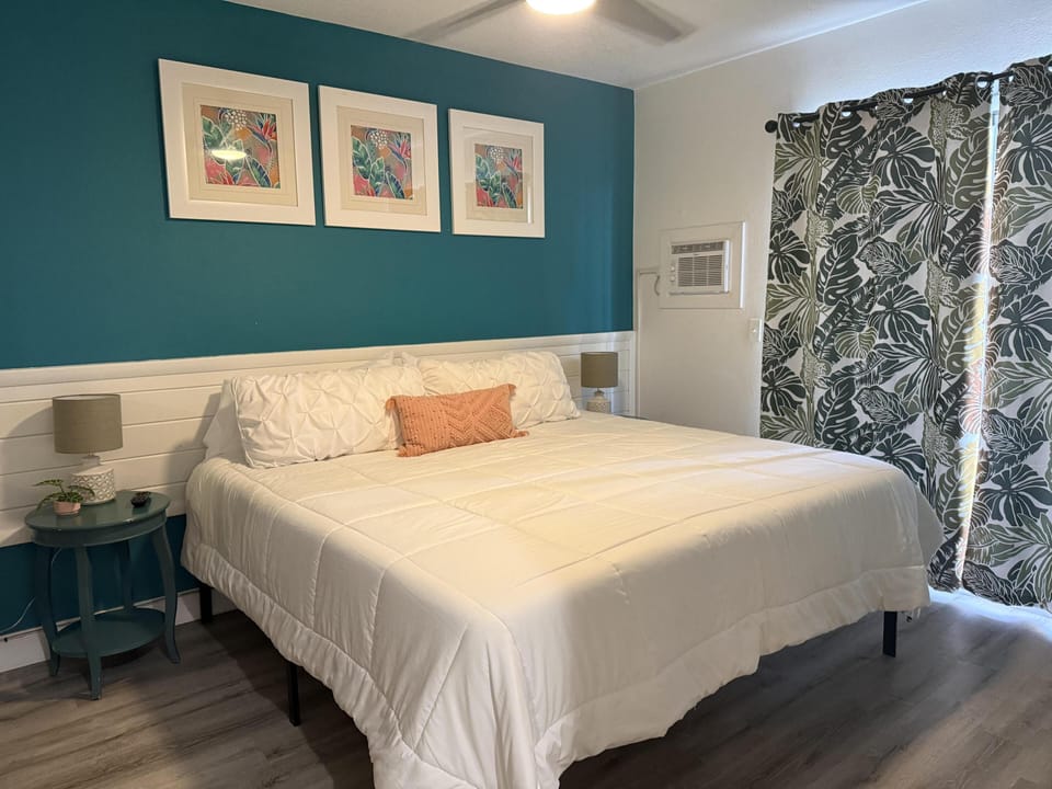 King-size bed with white quilt and coral pillow against teal accent wall with tropical artwork.