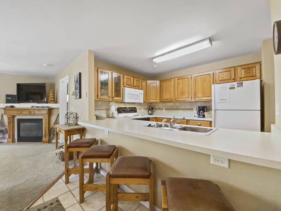 A kitchen with wooden cabinets, white appliances, and a countertop with stools. Living area in the background with a fireplace, TV, and small table.
