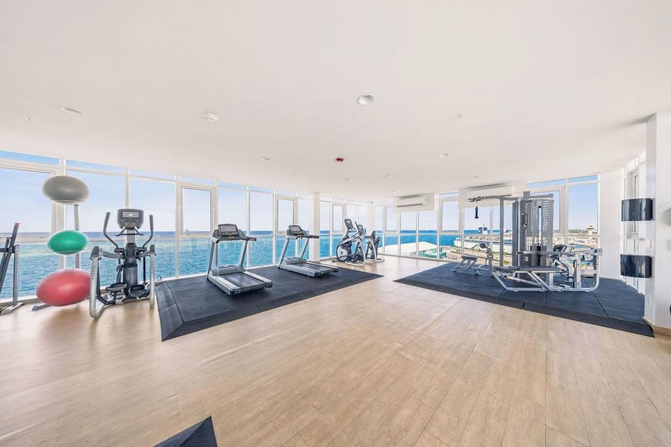 A modern fitness center spans floor-to-ceiling windows overlooking the sparkling sea, where treadmills, bikes, and weights await your waterfront workout.