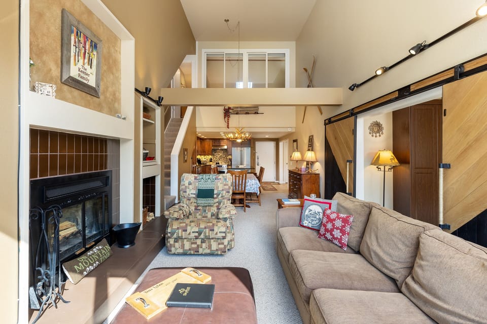 A cozy living room with a fireplace, brown sofa, patterned armchair, and a dining area visible in the background. The room features a high ceiling and decorative elements on the walls.