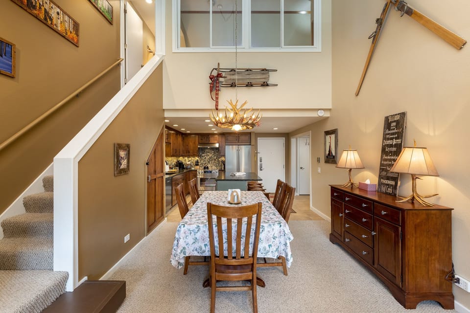 A dining area with a wooden table and six chairs, a floral tablecloth, a sideboard with lamps, and a staircase leading to an upper level. The space features rustic decor and an open layout.