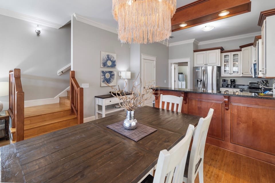Beautiful wood dining table beneath a stunning statement chandelier. Open-concept space flows into a fully equipped kitchen with stainless steel appliances, warm wood cabinetry, and plenty of counter space.