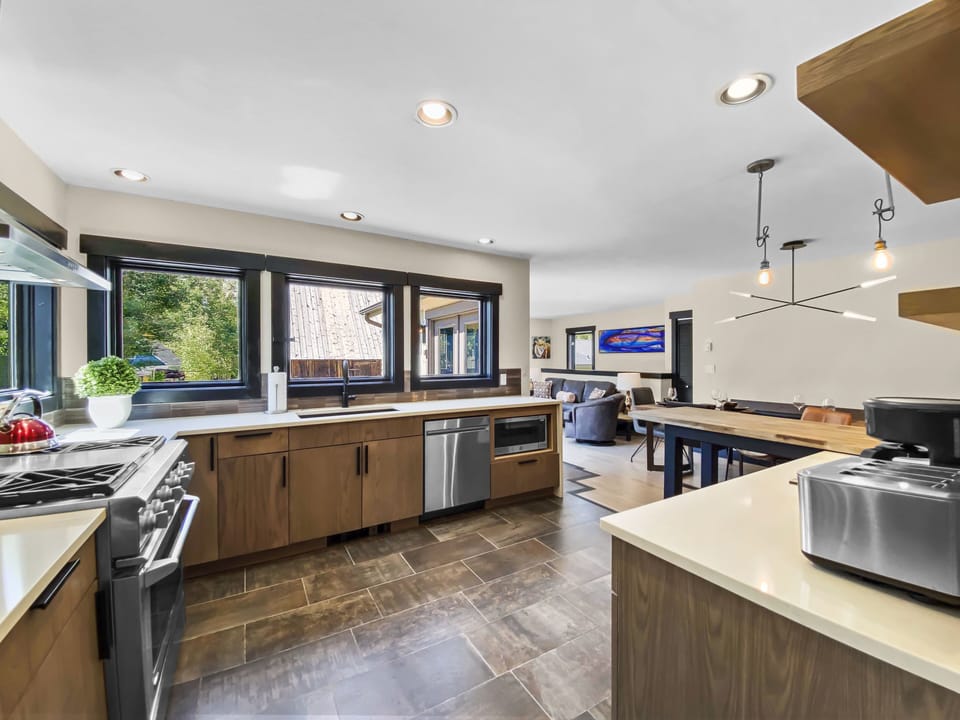 Modern kitchen with wooden cabinets, stainless steel appliances, and large windows. Dining area and living room with sofa and wall art visible in the background.