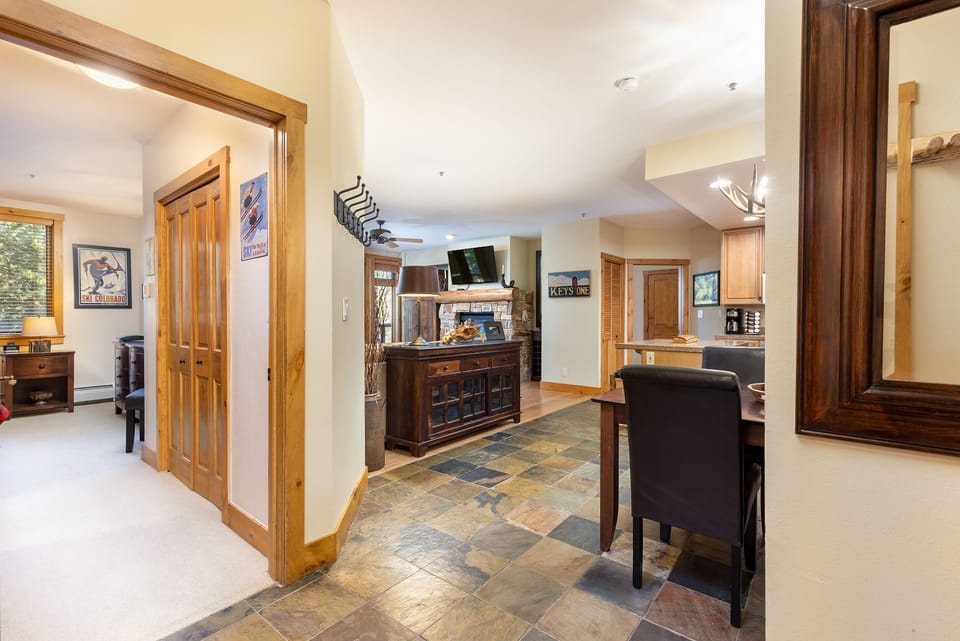View of a hallway leading to a kitchen and living area with stone-tile flooring. Wooden doors and trim are visible, along with a dining table, chairs, and a television in the background.
