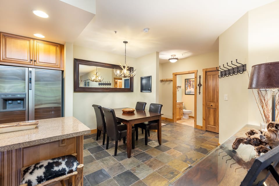 Modern kitchen and dining area featuring wood cabinetry, a large mirror, a rectangular dining table with four chairs, tiled flooring, and a view into a hallway leading to a small bathroom.