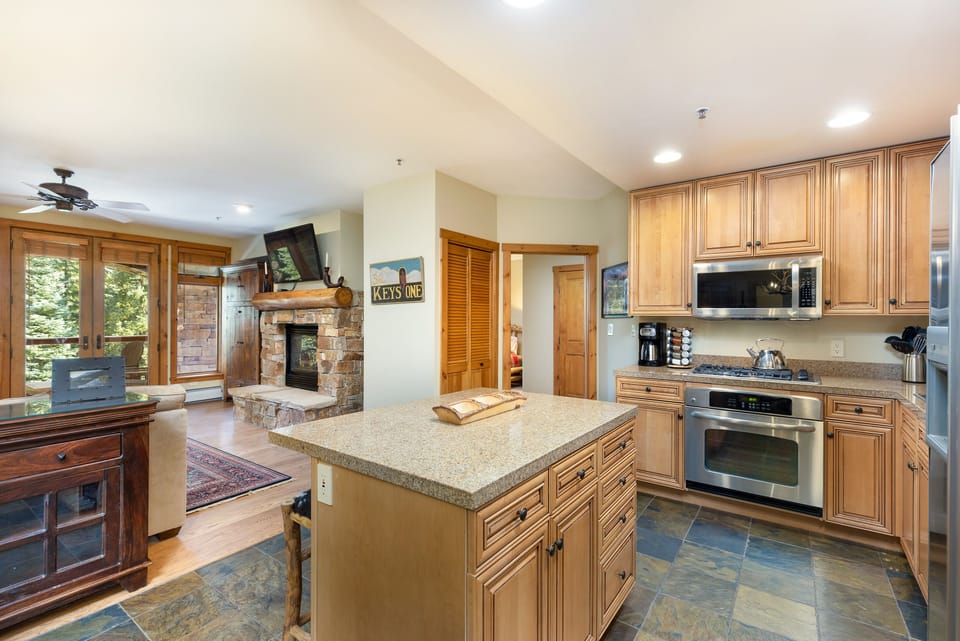 A modern kitchen with wooden cabinets, stainless steel appliances, and a central island. To the left is a cozy living area featuring a stone fireplace, wooden furniture, and large windows.