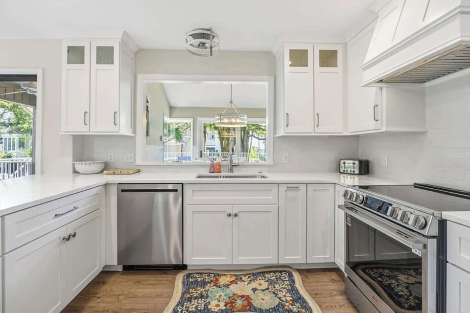 Bright cabinetry and stainless steel appliances create a clean, airy chef's space. A wide pass-through window connects the kitchen to the lush greenery outside, inviting the soft afternoon glow into every corner.