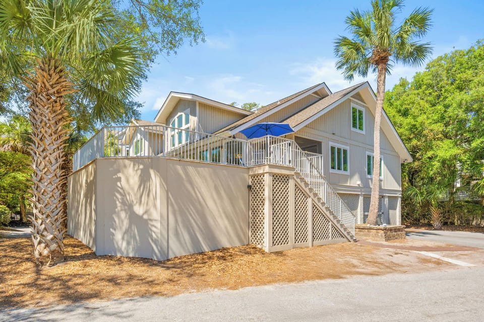 Palm trees frame this elevated beach home, where a wide wrap-around deck sits under the bright afternoon sky. Lattice detailing and soft tan siding create a breezy, laid-back atmosphere perfect for golden hour gatherings.