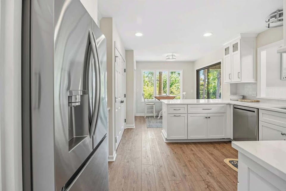 Stainless steel appliances and snowy cabinetry create a clean, modern chef’s space. Afternoon glow pours through the windows, highlighting the warm wood tones and making this airy kitchen the heart of the home.