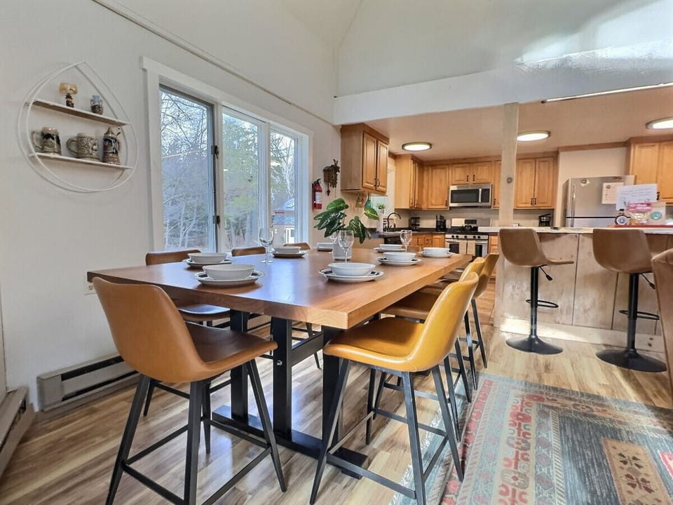 Bright dining area featuring a large wood table set for a group. It includes modern tan leather chairs and large windows that overlook a wooded outdoor view.