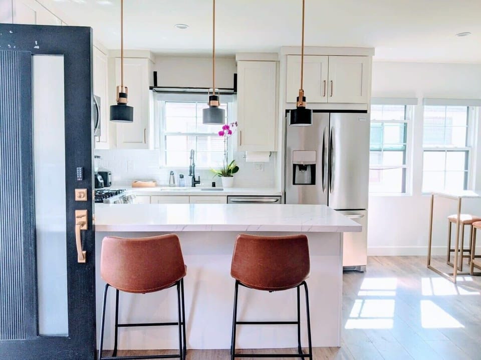 Welcome to our bright, modern kitchen featuring a clean white island with leather barstools and sleek pendant lighting that opens up into a sunlit living space.
