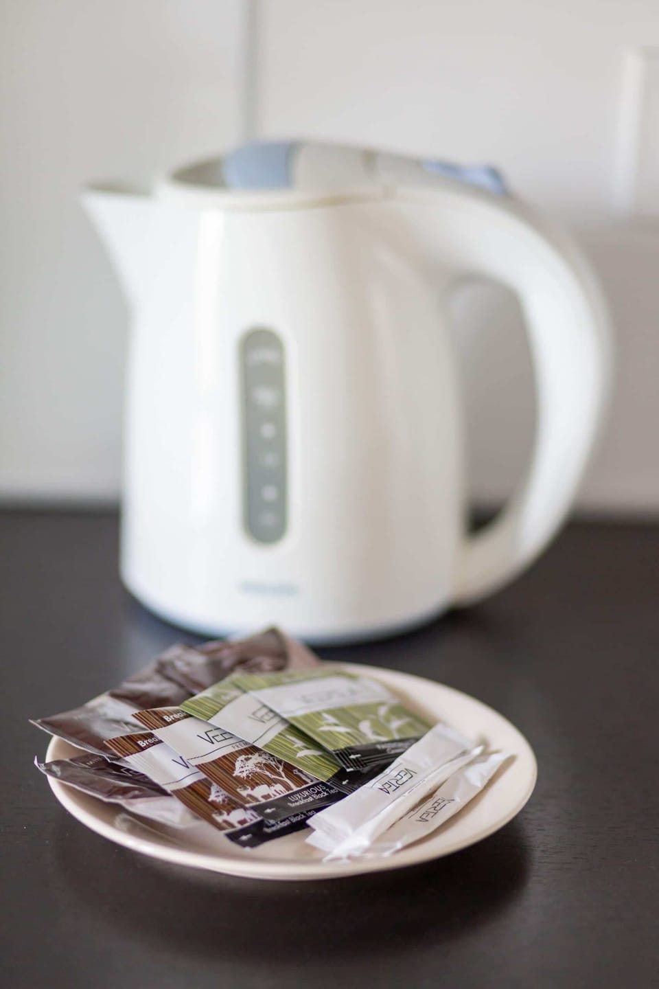 A close-up of a white electric kettle on a black tray, accompanied by tea and coffee supplies.