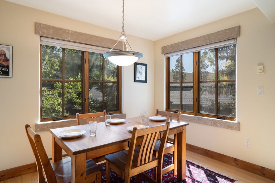 A dining area with a wooden table set for four, surrounded by wooden chairs. The room has two large windows with views of trees, and a ceiling light fixture hanging above the table.