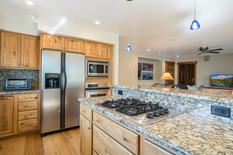 Modern kitchen with granite countertops, stainless steel appliances, a gas stove, and wooden cabinetry. Ceiling lights and a living area with a TV are visible in the background.