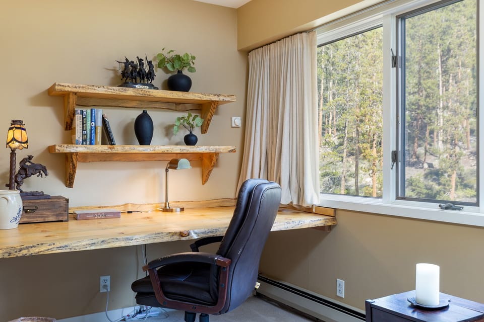 Cozy home office with wooden desk and shelves, books, vases, and decorative items. A large window overlooks a forest. A cushioned chair, table lamp, and candle complete the setup.
