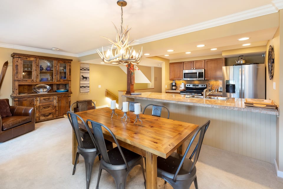A cozy dining and kitchen area features a wooden dining table with six metal chairs, a countertop with a breakfast bar, stainless steel appliances, and rustic decor including a chandelier and hutch.