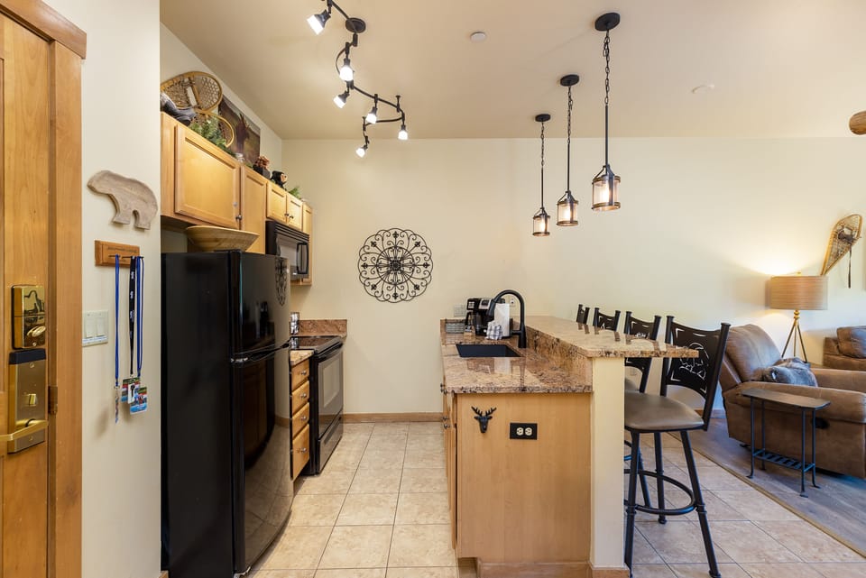 A modern kitchen with pendant lighting, granite countertops, black appliances, and wooden cabinets. To the right, a small bar area adjoins the living room with a couch and a lamp.