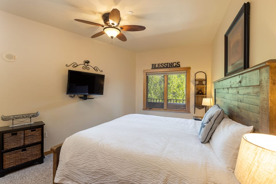 A cozy bedroom features a wooden bed with white bedding, a ceiling fan, a wall-mounted TV, wicker drawers, and a window with a view of trees. The word "Blessings" is displayed above the window.