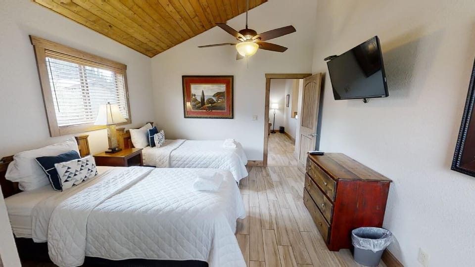 Your spacious bedroom features two plush beds with crisp linens, rustic wood-beam ceilings, a ceiling fan, and a wall-mounted TV—perfect for unwinding after a day of exploring.