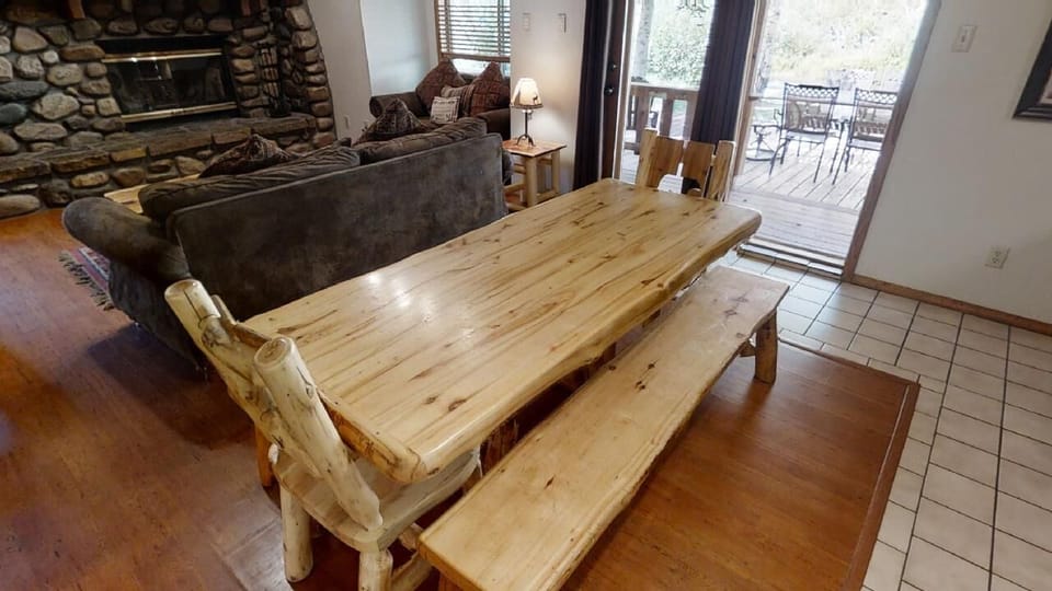 Step into your cozy mountain retreat featuring a rustic log dining table, plush seating by the stone fireplace, and your private deck overlooking natural surroundings—perfect for memorable meals and relaxation.