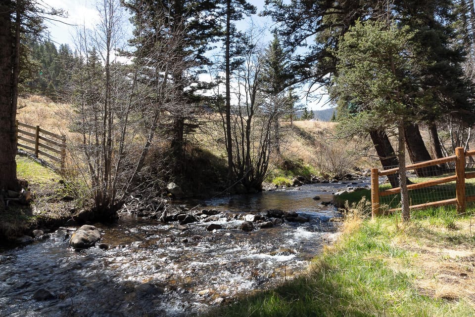 A peaceful stream winds through the wooded property grounds, bordered by rustic fencing and surrounded by tall pines and natural landscape—a serene backdrop for your retreat.