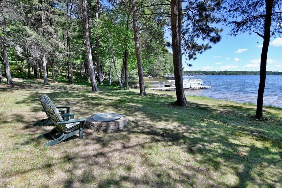 A grassy yard featuring a stone fire pit and wooden Adirondack chairs, leading down to a calm lake with a wooden dock under a clear blue sky.